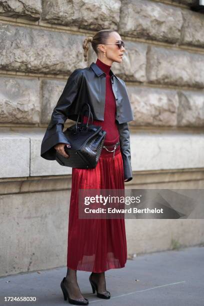 Viktoria Rader wears sunglasses, a burgundy turtleneck pullover, a black leather jacket, a large leather black crocodile pattern Hermes bag, a red...