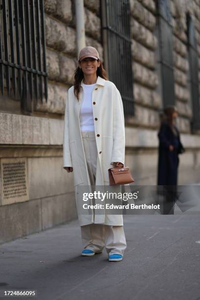 Guest wears a cap hat, a white t-shirt, a white long coat, flared wide-leg pants, blue shoes, outside Hermes, during the Womenswear Spring/Summer...