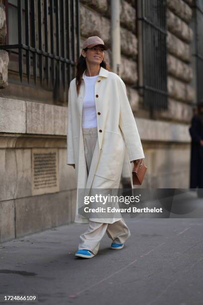 Guest wears a cap hat, a white t-shirt, a white long coat, flared wide-leg pants, blue shoes, outside Hermes, during the Womenswear Spring/Summer...