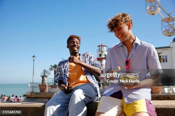 two young male adults sharing food together at the seaside - ocio fotografías e imágenes de stock