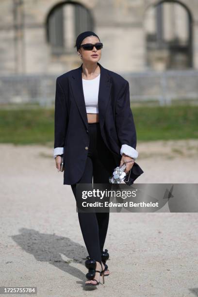 Guest wears a headband, sunglasses, a white crop top, a navy dark blue oversized blazer jacket, leggings, high heels shoes, outside Loewe, during the...