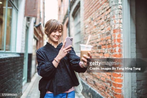 young pretty asian woman taking picture of her iced cold bubble tea with smartphone while walking through an alley in downtown district - equipment stock pictures, royalty-free photos & images