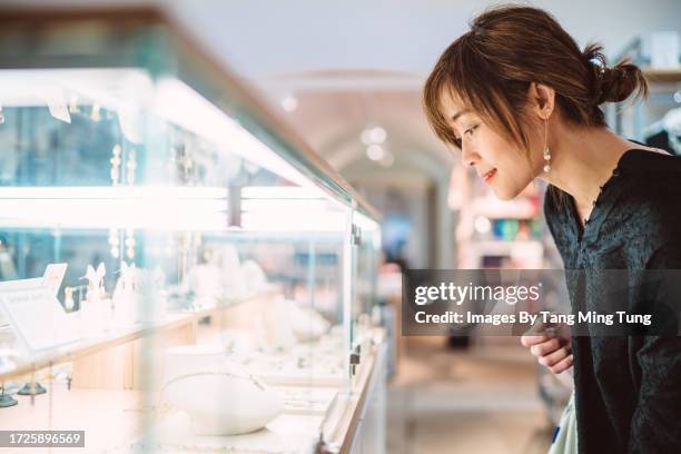 young pretty asian woman looking at jewellery through a glass display cabinet in a jewellery shop - jeweller stock pictures, royalty-free photos & images