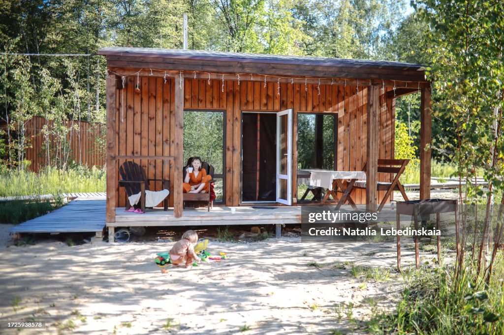 Children on trip to house in forest: teenager drinks cocoa on veranda and watches toddler in sandbox