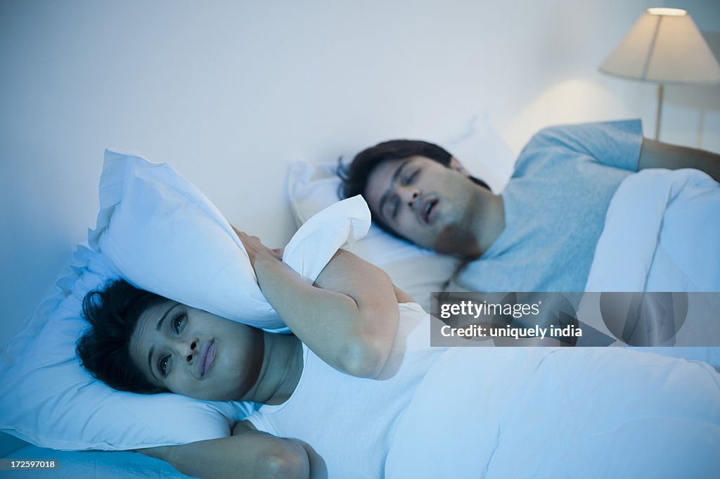 Man snoring with his wife covering ears with pillow on the bed