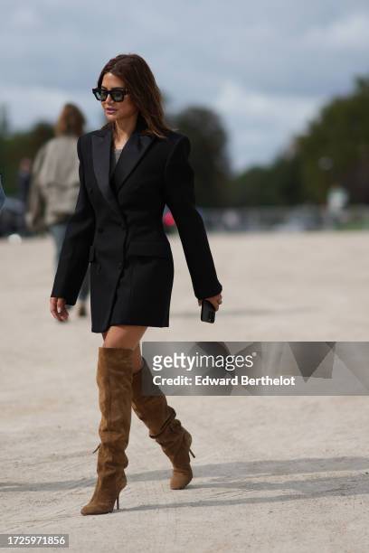 Christine Centenera wears sunglasses, a black oversized blazer jacket, knee high brown suede high heels boots, outside Loewe, during the Womenswear...