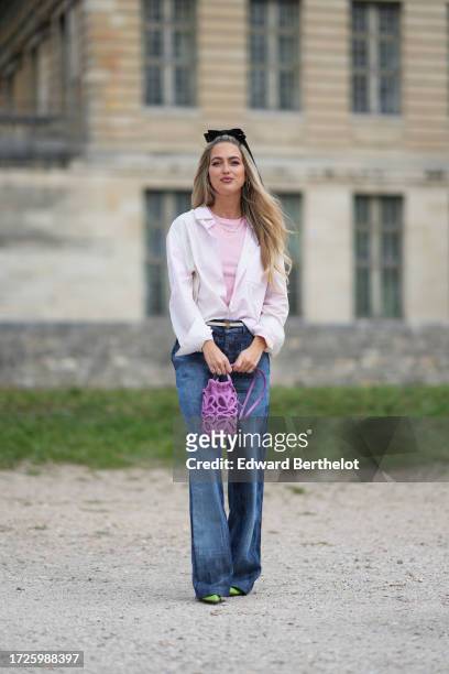 Emili Sindlev wears a bow tie headband, a pastel pink top, a white shirt, a necklace, a thin belt, blue denim flared jeans pants, a purple Loewe bag,...