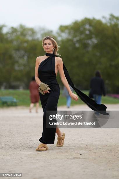 Xenia Adonts wears earrings, a black gathered long asymmetric flowing slit dress, a khaki gathered leather bag worn as a bracelet / cuff, brown...