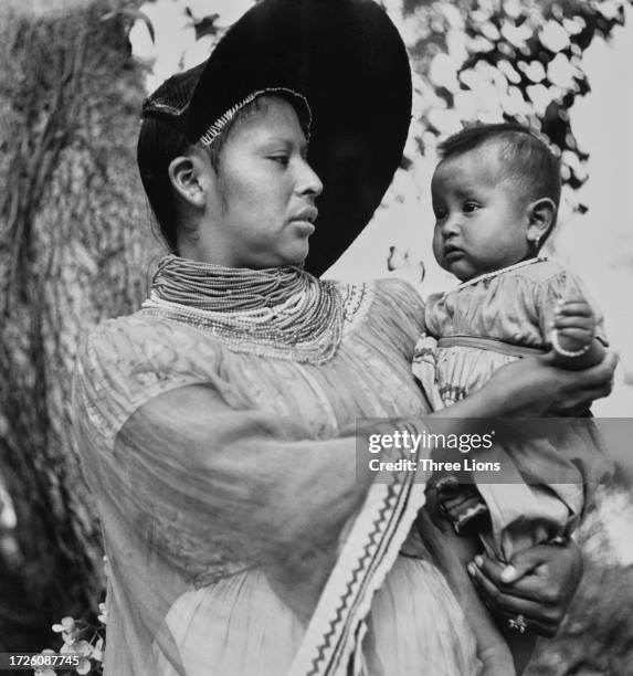 Native American Seminole woman in traditional clothing, with beaded necklaces and a hat with a wide brim to the front and side, holding her child in...