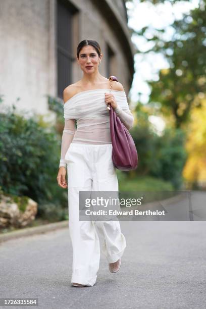 Belen Hostalet wears an off-shoulder white gathered tulle mesh top, white flared pants, a purple leather gathered bag, outside Christopher Esber,...