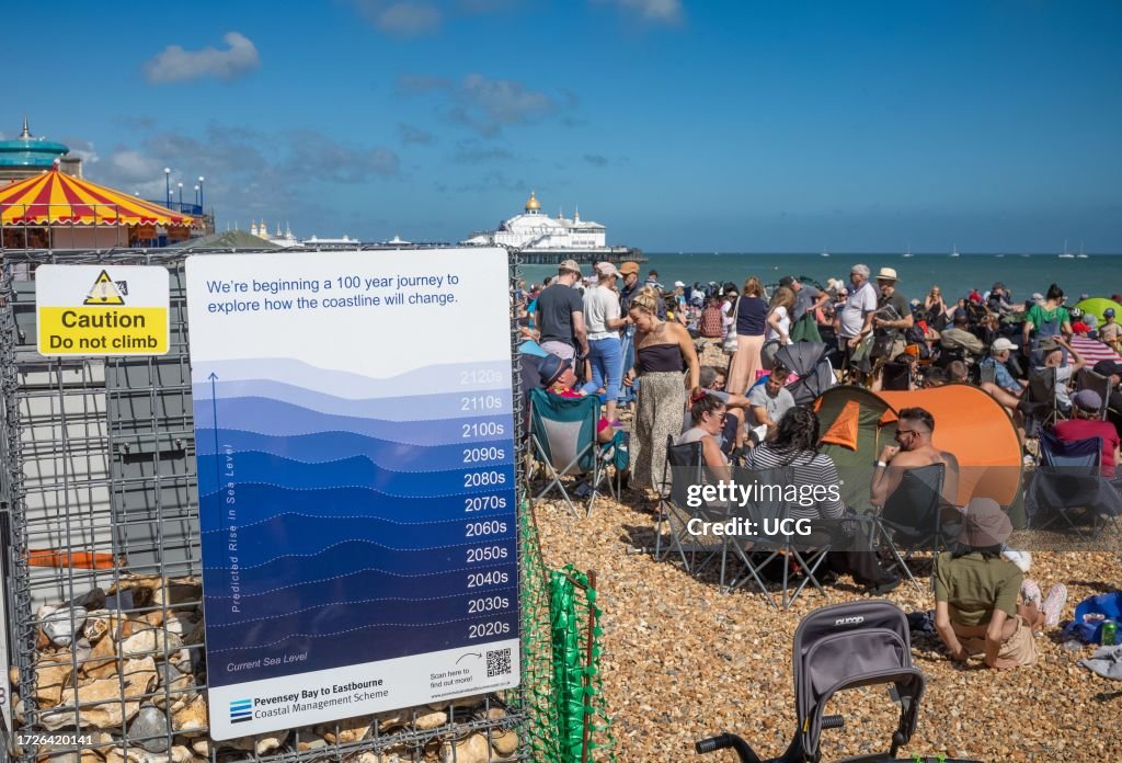 People on the beach next to a poster about rising sea levels in Eastbourne, East Sussex, UK.