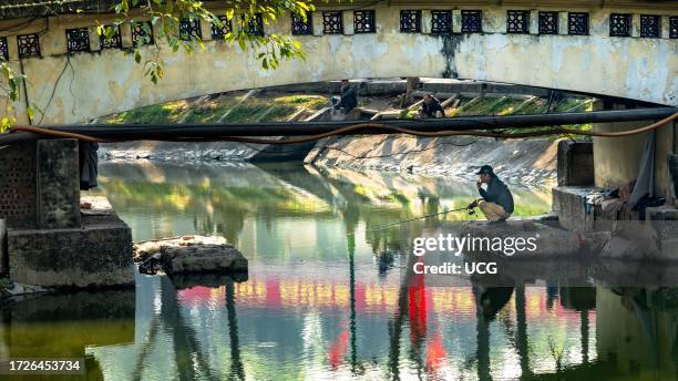 Vietnamese man smokes a cigarette as he fishes under a bridge on Thien Quang Lake in Hanoi, Vietnam. The red reflections are communist party...