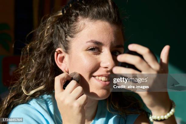 beautiful woman in casual clothes smiling sitting looking towards the camera while applying makeup on her cheek holding a mirror, frontal view - human face frontal stock pictures, royalty-free photos & images