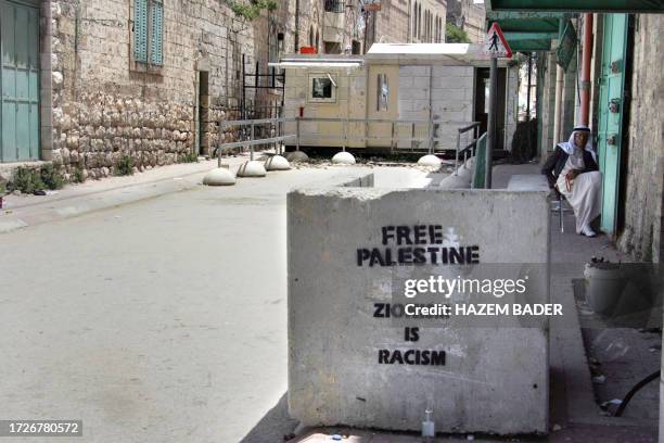 Palestinian man sits outside his store near an Israeli military checkpoint set-up across a street to control the movement of Palestinians and others...