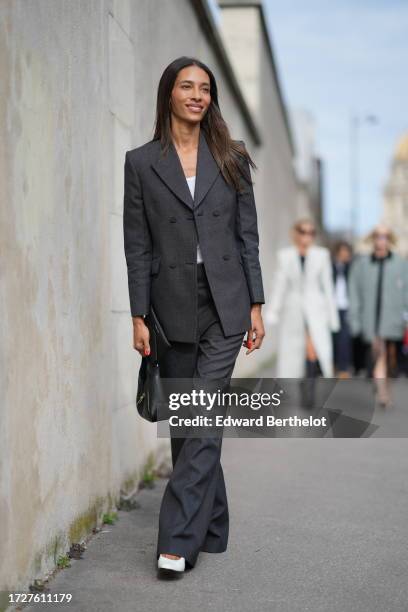 Guest wears a white top, a dark gray blazer jacket, flared suit pants, a black leather bag, white pointed shoes, outside Givenchy, during the...