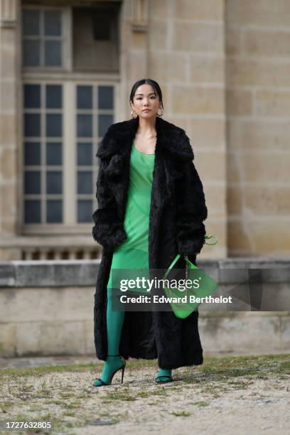 Yoyo Cao wears large golden earrings, a black faux fur fluffy long winter coat, a low-neck green dress, dark green tights, a green leather Givenchy...