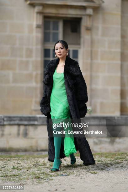 Yoyo Cao wears large golden earrings, a black faux fur fluffy long winter coat, a low-neck green dress, dark green tights, a green leather Givenchy...