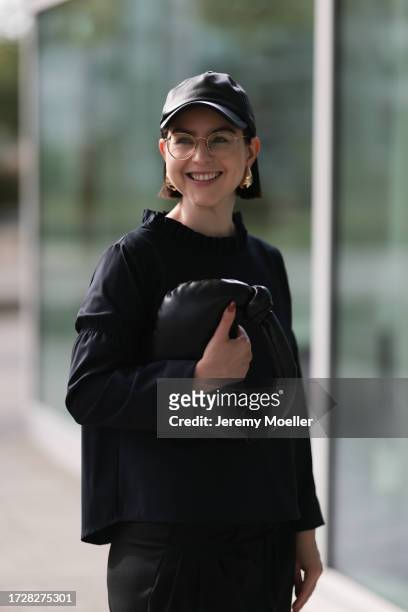 Maria Barteczko is seen wearing a black leather baseball cap from Arket, round retro glasses with golden frame from Ray-Ban, golden logo earrings...