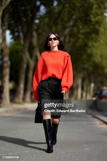 Guest wears sunglasses, golden earrings, an orange ribbed wool oversized tucked in pullover, black shorts, knee high leather boots, outside Chloe,...