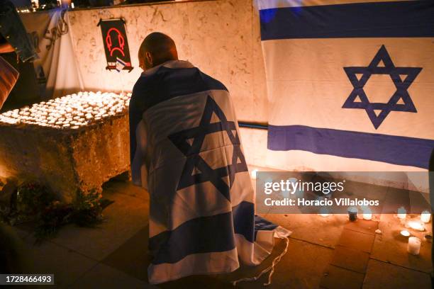 Participant draped in an Israeli flag kneels before lit candles during a vigil organized by the Lisbon Jewish Community for Israeli victims of the...
