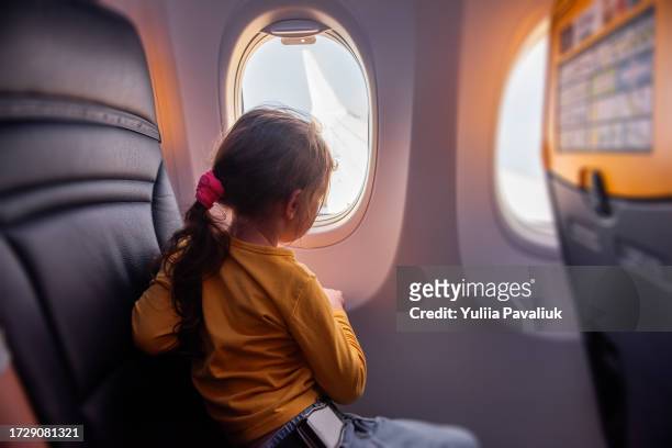 little girl looks out window porthole of airplane at wing. child is not afraid to fly on plane - travel stock-fotos und bilder
