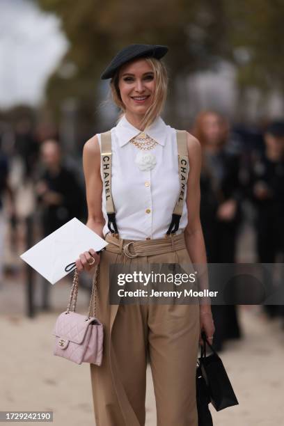 Guest is seen outside Chanel show wearing black barett hat, white Chanel blouse with white camellia detail, golden necklaces, Chanel logo suspenders,...