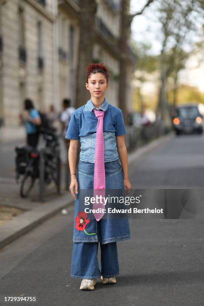 Caroline Takrarit wears a blue denim long dress with a red printed flower / integrated corset, a long pink tie, flared jeans, sneakers, outside...