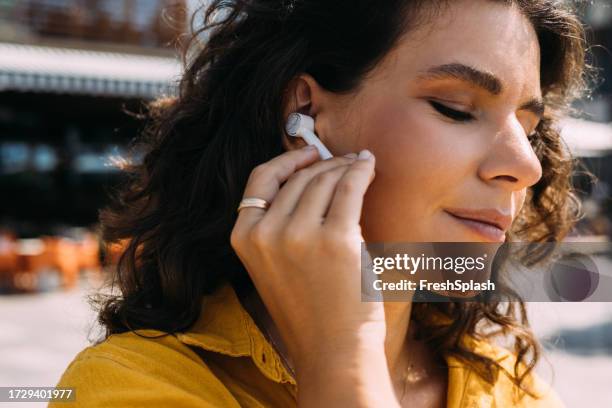 a happy beautiful woman with curly hair using her earbuds while standing on the street - in ear headphones stock pictures, royalty-free photos & images