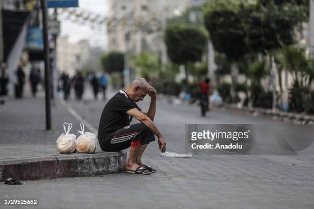 Palestinian man sits on a sidewalk after shopping from a street vendor following the suspension of food, water and electricity by Israeli authorities...