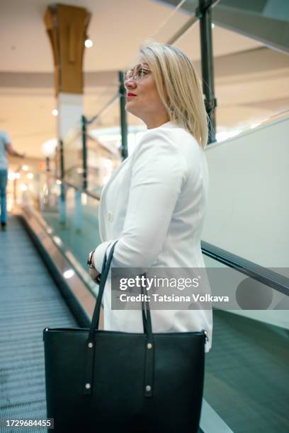 stylish blonde woman in white blazer immerses herself in lively ambiance of contemporary shopping center while riding escalator - ambiance girly stock pictures, royalty-free photos & images