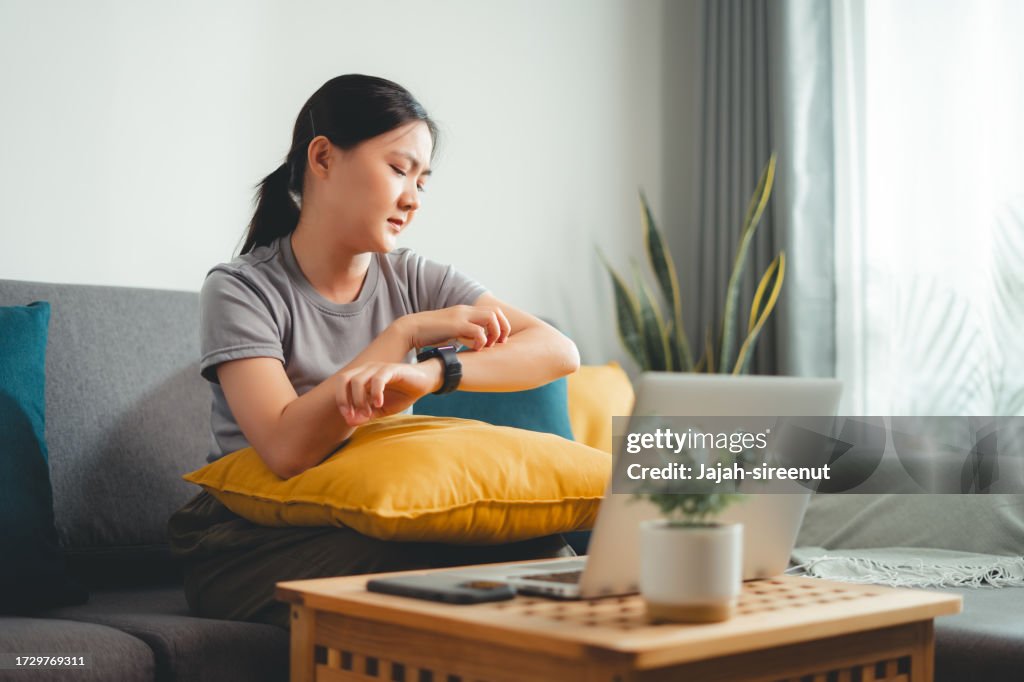 Asian woman irritate itching dry skin scratching her skin sitting on sofa in living room at home.