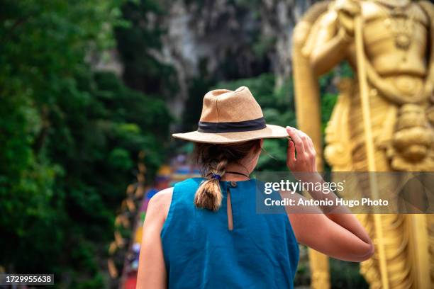 young woman admiring the batu caves in malaysia while touching her hat with one hand. - thaipusam stock pictures, royalty-free photos & images