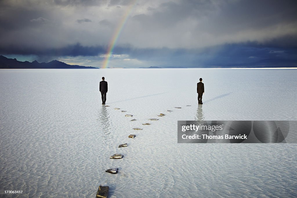Two men on forked pathway in water under rainbow
