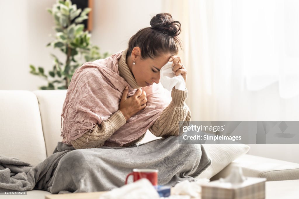 Very sick young woman sitting on a couch holding her forearm and handkerchief at her nose fighting fever.