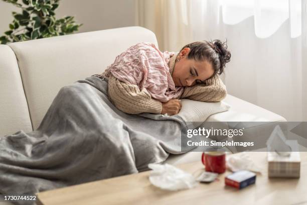 very sick young woman slipping on a couch holding her forearm and handkerchief at her nose fighting fever. - winterdepression stock-fotos und bilder