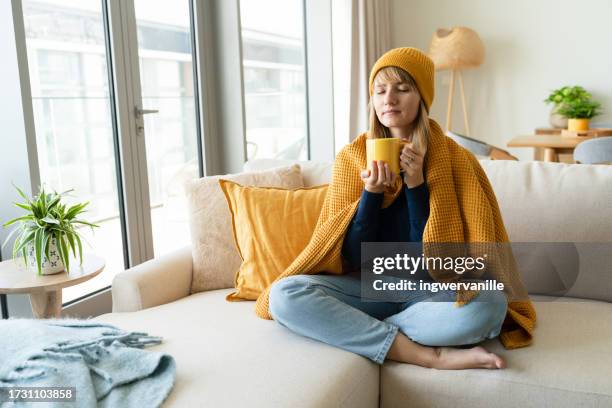 woman in woolen hat wrapped in blanket drinking tea in the living room - virus grippal photos et images de collection