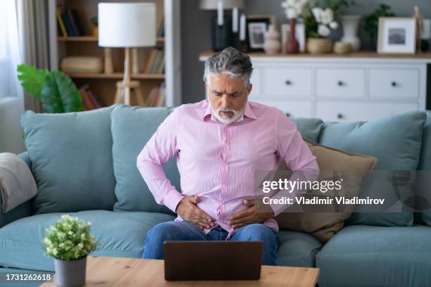 un uomo che soffre di mal di stomaco mentre è seduto sul divano di casa - colite foto e immagini stock