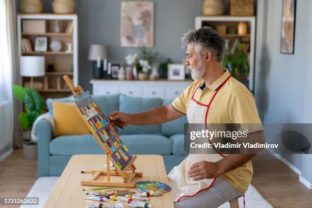 a mature man is painting a picture on an easel in a modern living room - art therapy stock pictures, royalty-free photos & images