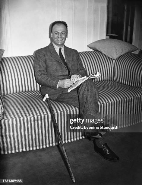 French fashion designer Pierre Balmain seated on a sofa with a magazine and a walking stick, circa 1950.