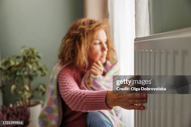 a woman wrapped in a blanket trying to keep warm by the heating radiator - domestic room stock pictures, royalty-free photos & images
