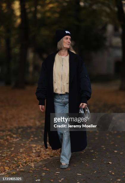 Victoria Thomas is seen wearing a dark blue beanie from Hernameis the label, a golden necklace, a long black coat with bomber jacket sleeves, a beige...