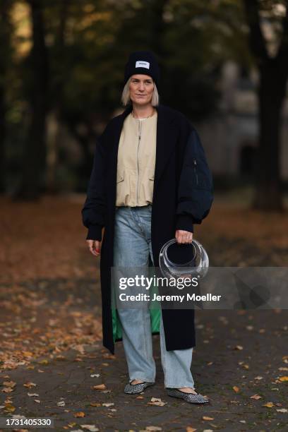 Victoria Thomas is seen wearing a dark blue beanie from Hernameis the label, a golden necklace, a long black coat with bomber jacket sleeves, a beige...