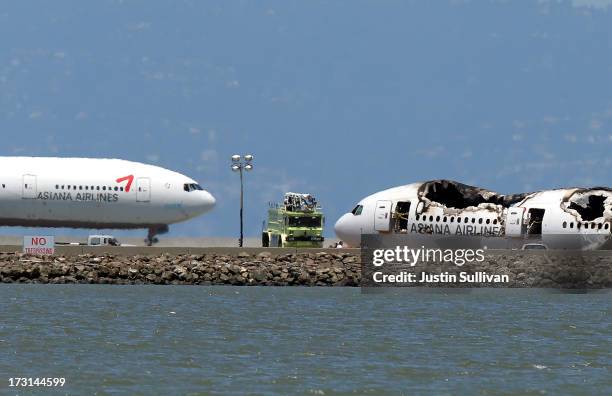 An Asiana Airlines flight enroute to South Korea, a Boeing 777, taxis by the wreckage of Asiana Airlines flight 214 as it sits on runway 28L at San...