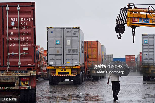 Man with an umbrella walks past freight trucks loaded with shipping containers at a freight depot near Nhava Sheva Port in Navi Mumbai, India, on...