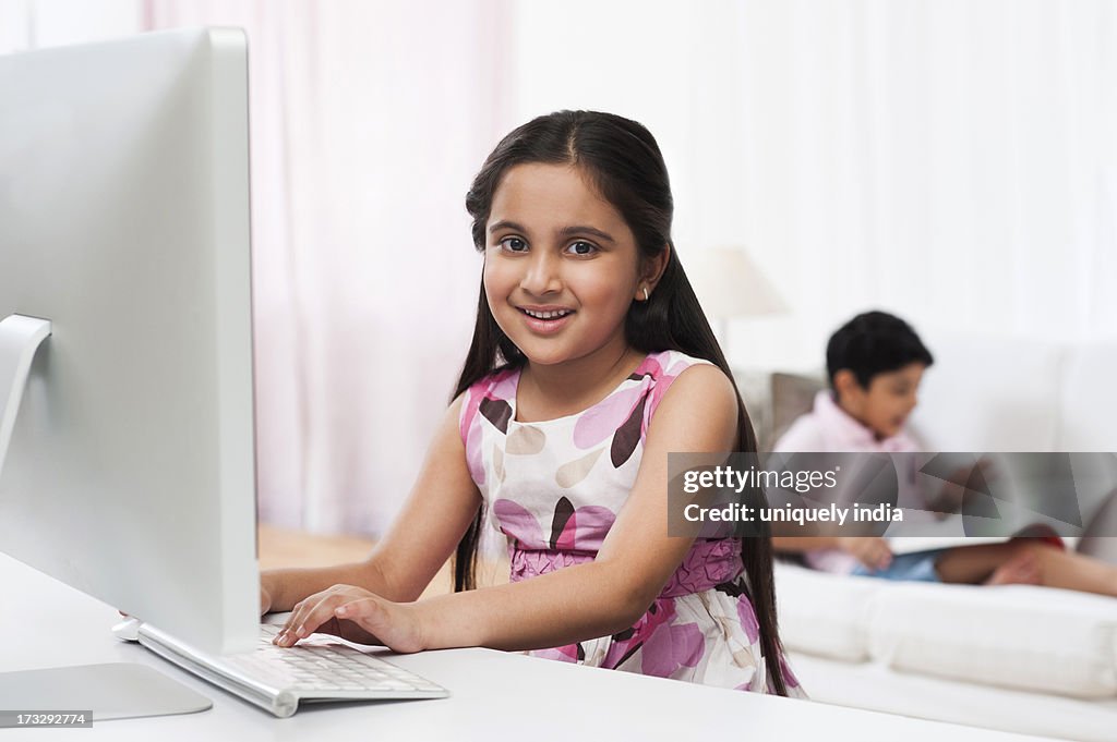 Girl using a computer with her brother reading a book in the background