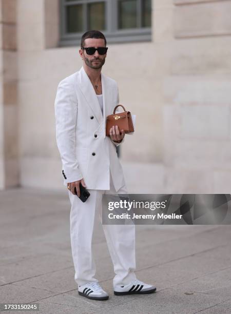 Guest seen wearing Ray-Ban black sunglasses, Hermès mini kelly brown bag, a white shirt, a white suit jacket and matching white suit pants and Adidas...