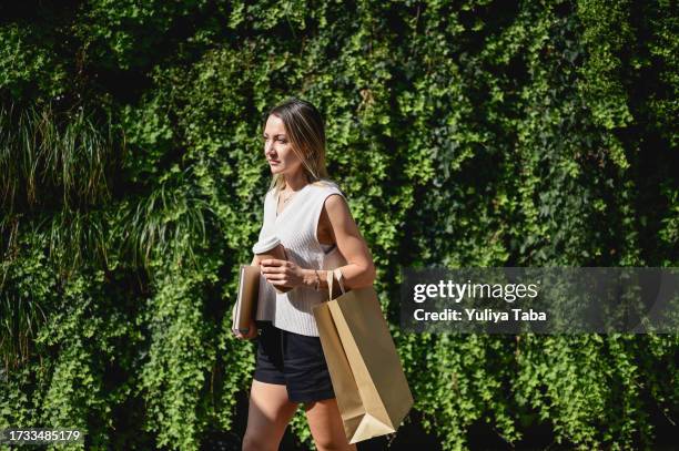 donna alla moda per adulti medi di fronte a un giardino verticale che tiene una tazza di caffè usa e getta e un sacchetto di carta. - disposable cup foto e immagini stock