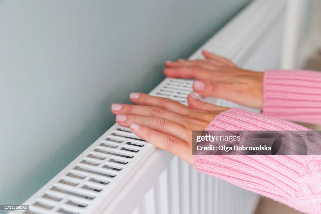 Close-up of a woman hands in a pink jumper keeping warm by a heating radiator