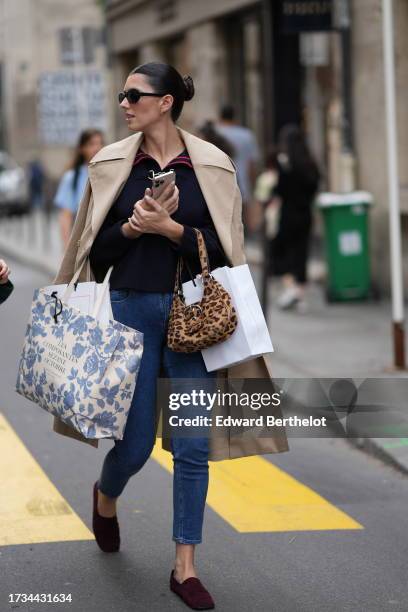 Guest wears sunglasses, a beige trench coat, a pullover, blue cropped jeans, purple suede shoes, a brown leopard print bag, outside Cecilie Bahnsen,...