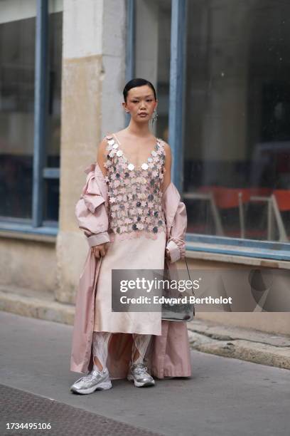 Guest wears a shiny silver sequined top over a lustrous pink dress, a pink coat, a silver bag, sneakers, outside Cecilie Bahnsen, during the...